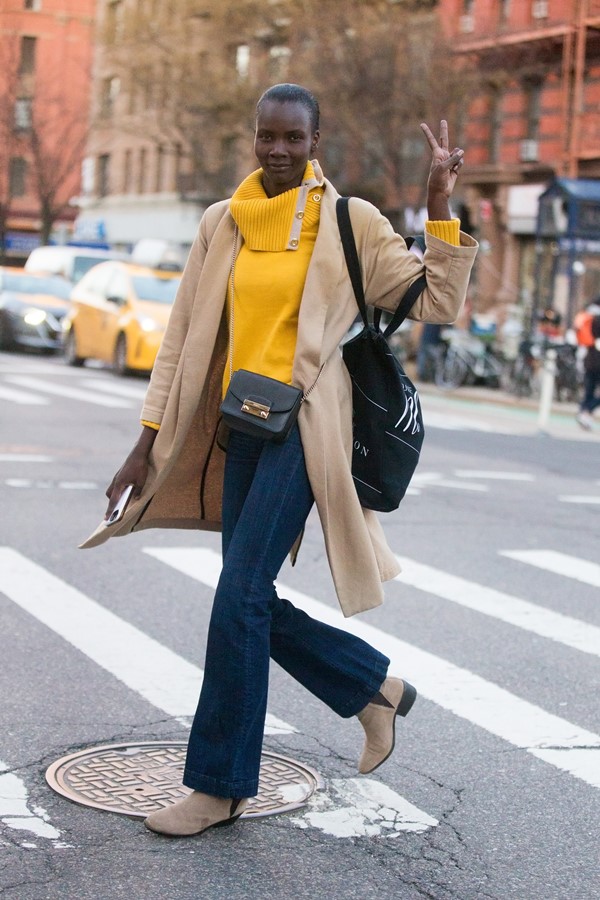 Mulher negra com cabelo raspado atravessando a faixa de pedestres. Ela usa uma calça jeans, casaco amarelo, sobretudo preto e duas bolsas pretas.