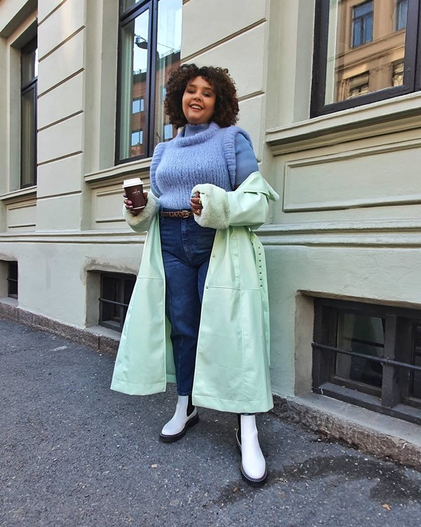 Mulher negra com cabelo curto, castanho e cacheado. Ela posa para foto na rua usando calça jeans azul, bota branca, tricot lilás e sobretudo verde claro.