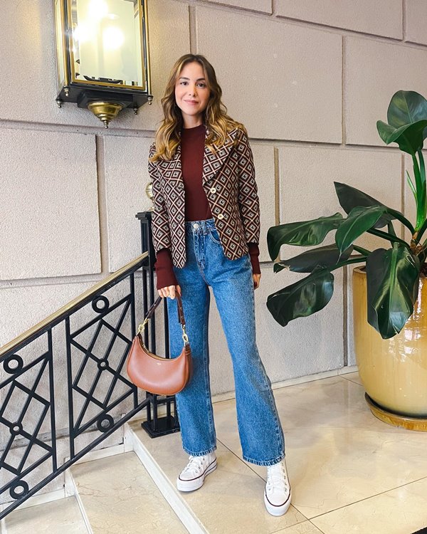 Mulher branca com cabelo louro, ondulado e curto. Ela posa para foto em uma escada e usa calça jeans azul, blusa, blazer e bolsa marrom.