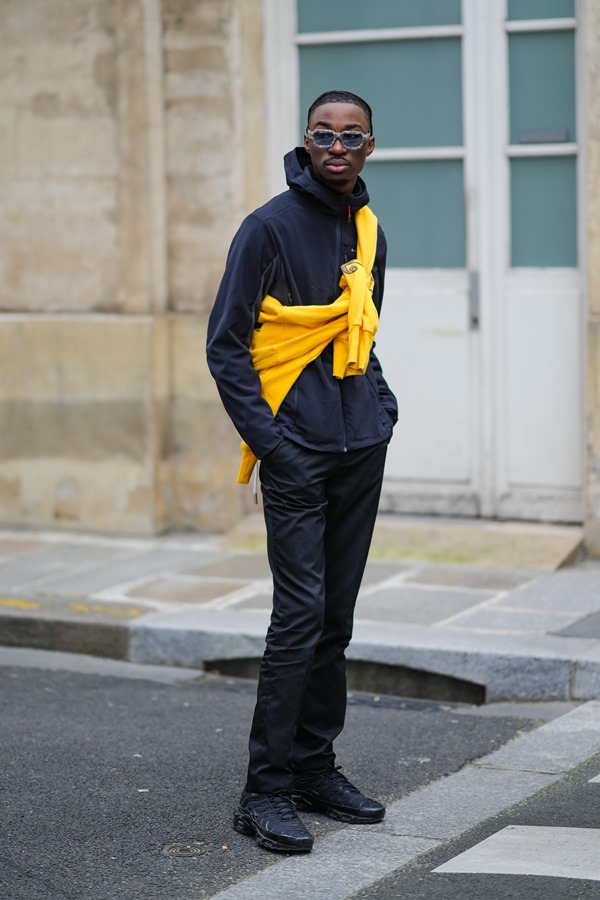 Homem negro de cabelo raspado posando para foto no street style. Ele usa roupa toda preta, tênis Air Max da Nike e um casaco amarelo amarrado.