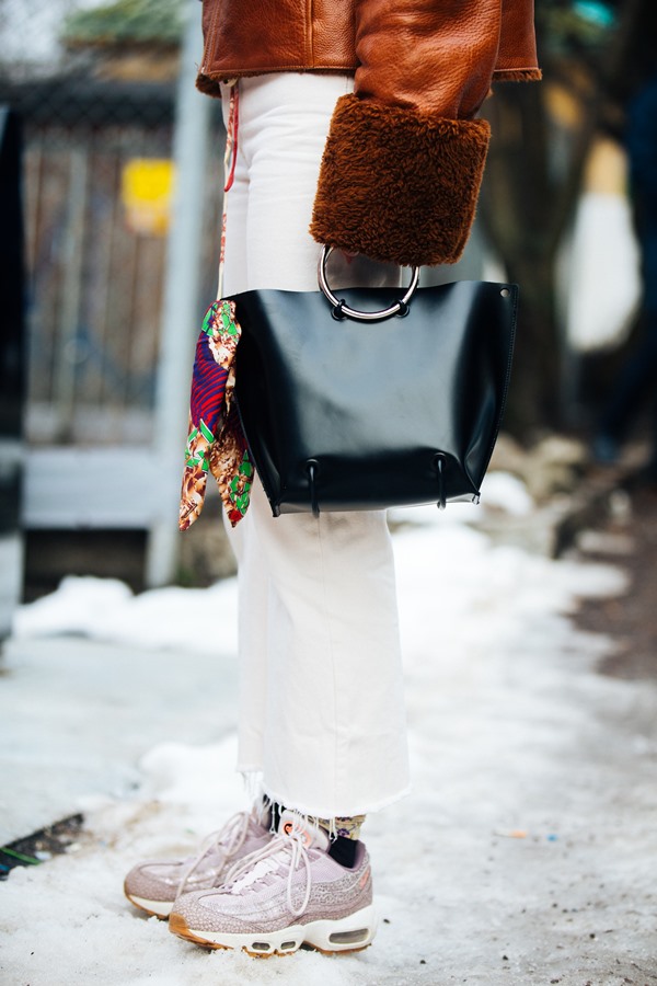 Detalhes da roupa de uma pessoa. Ela está em uma rua com neve e usa meia, tênis, calça, casaco e uma bolsa de couro.