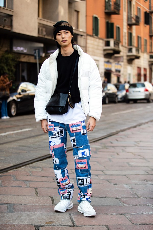 Homem asiático posando para foto de street syle. Ele usa uma calça jeans larga estampada, blusa preta e casaco branco, além de tênis Air Max brancos da Nike