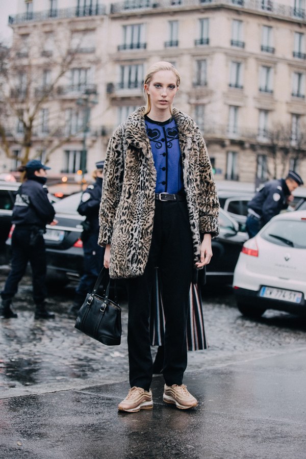 Mulher branca e loura, com cabelo longos penteados para trás, posando para foto de street style. Ela usa uma calça preta, tênis rose gold Air Max da Nike, blusa azul e casaco de onça.