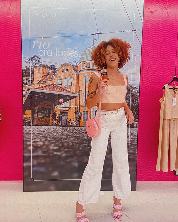 Mulher negra com cabelo cacheado curto e em tom de ruivo posa para foto em frente a painel da loja Shein no Rio de Janeiro. Ela usa calça branca, top, bolsa e salto alto rosa