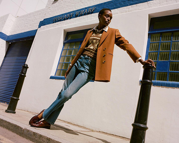 Mulher negra de cabelo raspado. Ela está na rua, se apoiando em um hudrante, e usa calça jeans, blusa, sapato e blazer marrom. As peças são da marca da Alexa Chung.