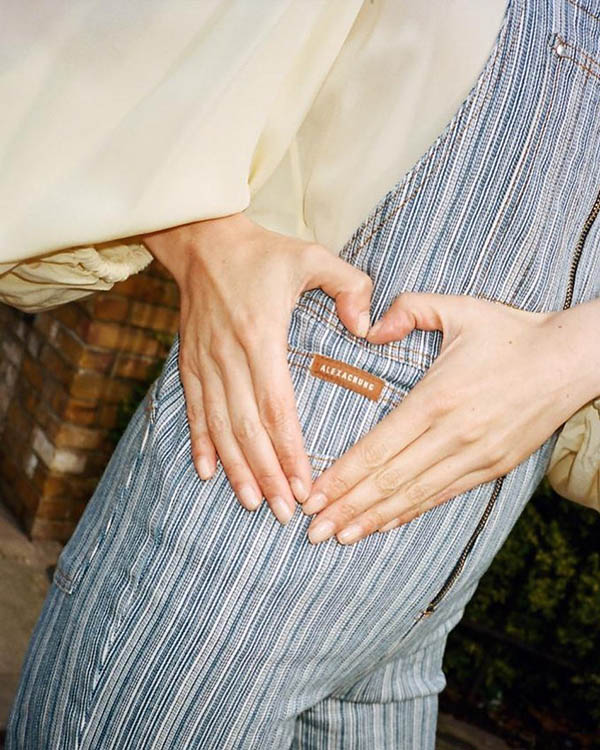 Pessoa vestindo um macacão jeans e msotrando a etiqueta da roupa. A peça é da marca da Alexa Chung.