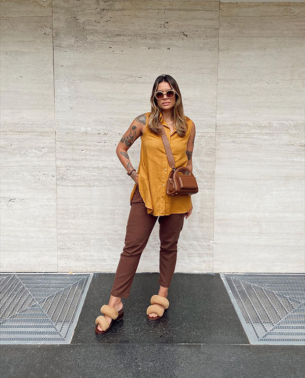 Mulher branca com cabelo louro e liso posando para foto em parede na rua. Ela usa calça e bolsa marrom, sandália rasteira bege e blusa mostarda.