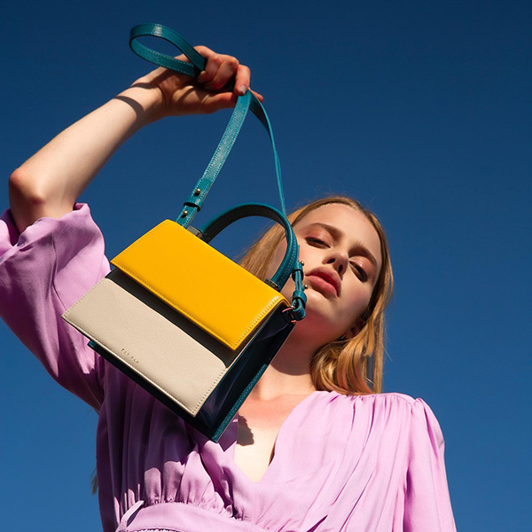 Mulher branca com cabelo liso e louro segurando uma bolsa. O acessório, que é da marca Parpar, possui alças azuis e o couro bege e amarelo. A modelo usa um vestido lilás.