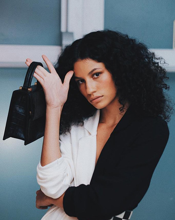 Mulher negra com cabelo cacheado preto segurando uma bolsa preta da marca Parpar. Ela está usando uma camisa bicolor preta e branca.