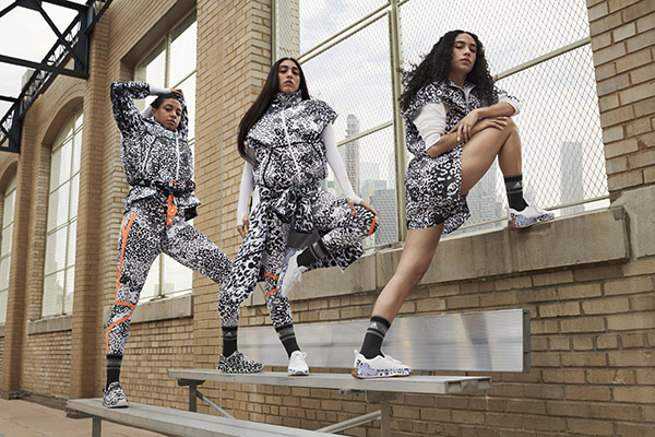 Três mulheres se alongam em uma escada de arquibancada de estádio para se exercitar. Ela usam roupas esportivas nas cores preta e branca da marca Adidas x Stella McCartney.