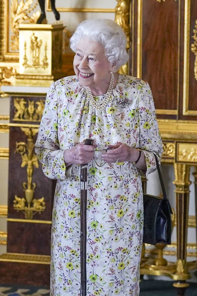 Foto colorida. Rainha Elizabeth com vestido florido, pérolas e uma bengala