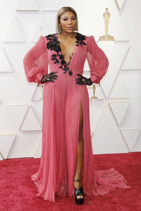 A tenista Serena Williams no tapete vermelho do Oscar. Ela usa um vestido rosa da marca Gucci, com luvas pretas. Ela está como cabelo curto liso e louro.