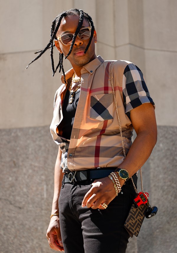 Homem negro de tranças fotografado na rua. Ele usa uma camiseta xadrez e um colete marrom, também xadrez, da marca Burberry. Ele está de calça preta e com uma bolsa de corrente da Louis Vuitton.