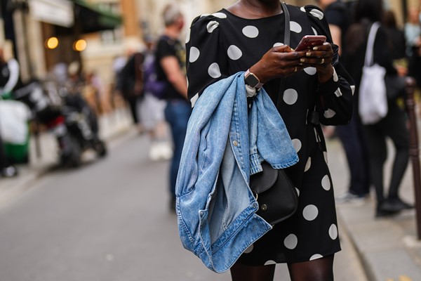 Mulher negra mexendo no celular e andando na rua. Na foto não dá para ver seu rosto. Ela veste um vestido preto de bolinha branca e uma jaqueta jeans.