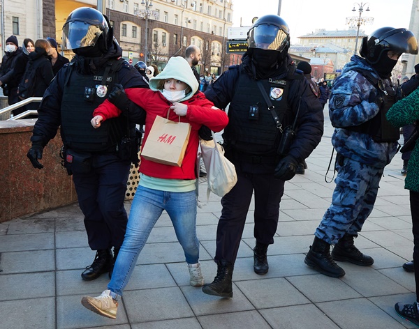 Mulher sendo escoltada por policiais em protestos na Rússia contra a invasão à Ucrânia. São dois policiais e cada um a segura por um dos braços. Ela usa um casaco vermelho e segura uma sacola marrom da marca H&M.