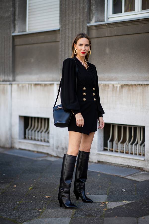 Mulher branca usa vestido que remete a blazer, com botões dourados frontais e mangas felpudas. Ela também está com bota preta acima do joelho