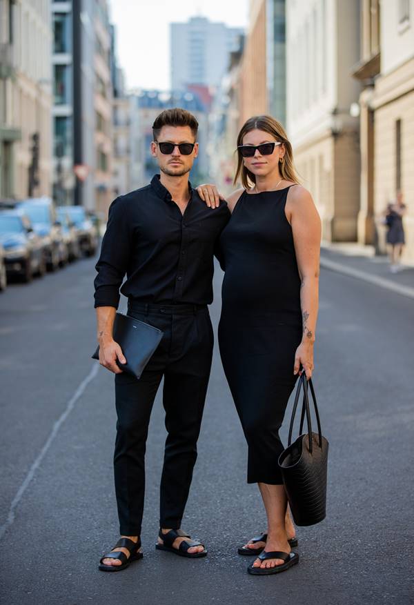 Casal usa looks pretos. Ele com camisa e calça de alfaitaria. Ela com vestido e bolsa