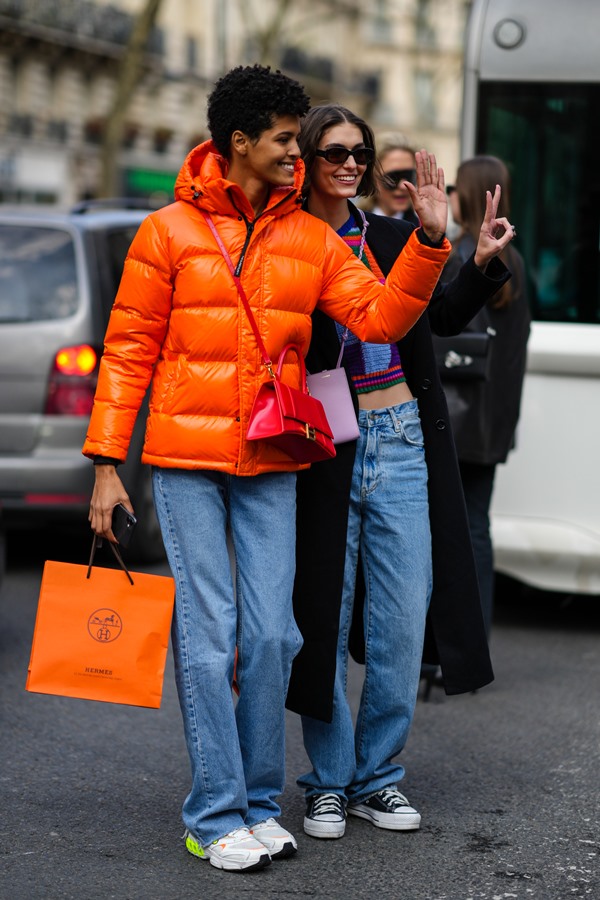 Modelos andando pela rua durante a Semana de Moda de Paris. A primeira é uma mulher negra de cabelo curto que usa calça jeans, casaco puffer laranja, uma bolsa rosa da Balenciaga e sacola laranja da marca Hermès. A mulher de trás é branca e loura e usa calça jeans e uma blusa cropped. As duas estão acenando com a mão