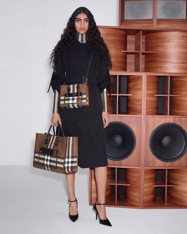 Modelo negra com cabelo longo e ondulado. Ela usa uma roupa preta e pochete e bolsa em estampa xadrez da marca Burberry.