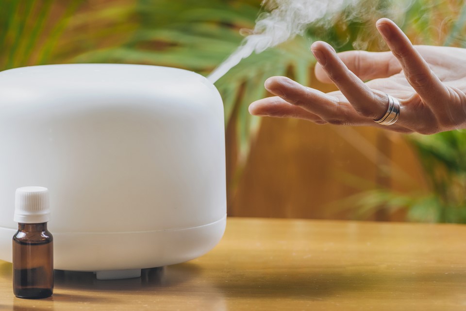 Mão toca o vapor que sai de difusor de aromas branco em mesa, ao lado de frasco com essência - Metrópoles