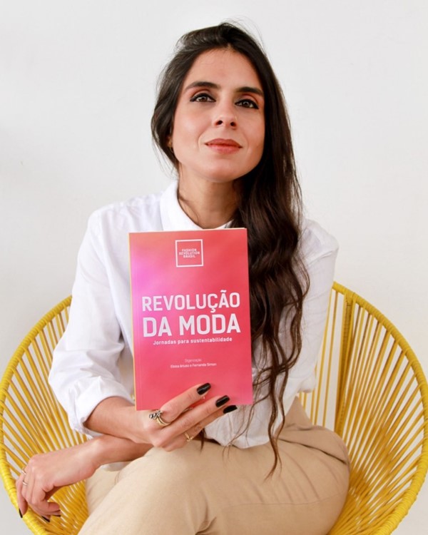 Mulher branca e com cabelos cumpridos castanhos e ondulados. Seu nome é Fernanda Simon. Ela é diretora do órgão Fashion Revolution Brasil. Na foto, está sentada em uma cadeira e usa calça bege e camisa branca. Ela segura um panfleto rosa onde é possível ler a frase "Revolução da Moda".