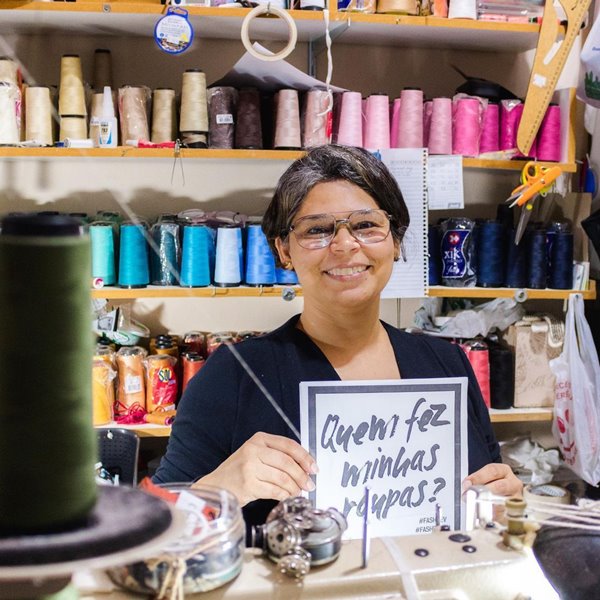 Mulher morena de cabelo liso curto e de óculos de grau. Ela segura uma placa com os dizeres "quem faz as minhas roupas?" e está sentada em uma mesa com máquina de costura. No fundo é possível ver rolos de linha de diferentes cores - Metrópoles