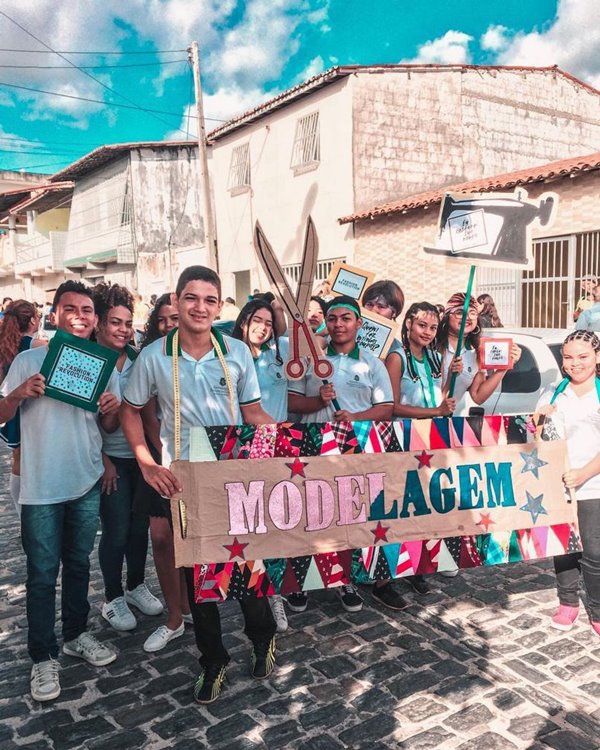 Estudantes do curso de moda uniformizados segurando uma placa escrita "modelagem". A foto é do bastidor de uma aula ensinando etapas de confecção das roupas