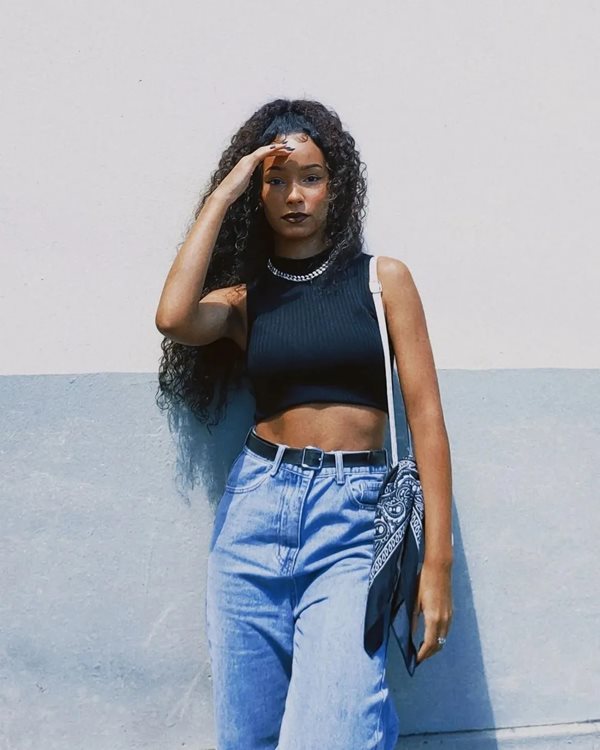 Mulher negra com cabelo cacheado longo encostada em muro, posando para foto. Ela está com um top preto, calça jeans, cinto preto e uma bolsa a tiracolo. As peças são da Shein.