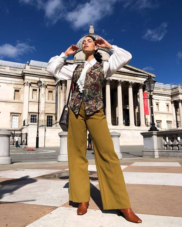 Mulher branca posa em frente a monumento usando calça vintage, colete estampado e camisa branca 