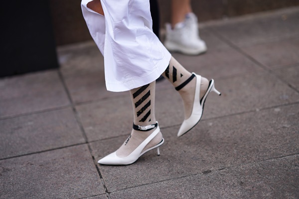 Mulher usando meias listradas esbranquiçadas, sapatos pontudos brancos e calça folgada, também branca, durante a Milan Fashion Week Outono/Inverno