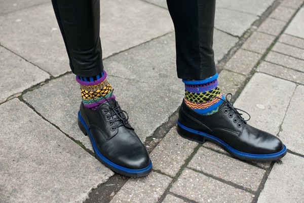 Homem usando sapatos pretos. Na foto é possível ver apenas seus pés. Além do calçado, ele usa meia colorida e calça preta.