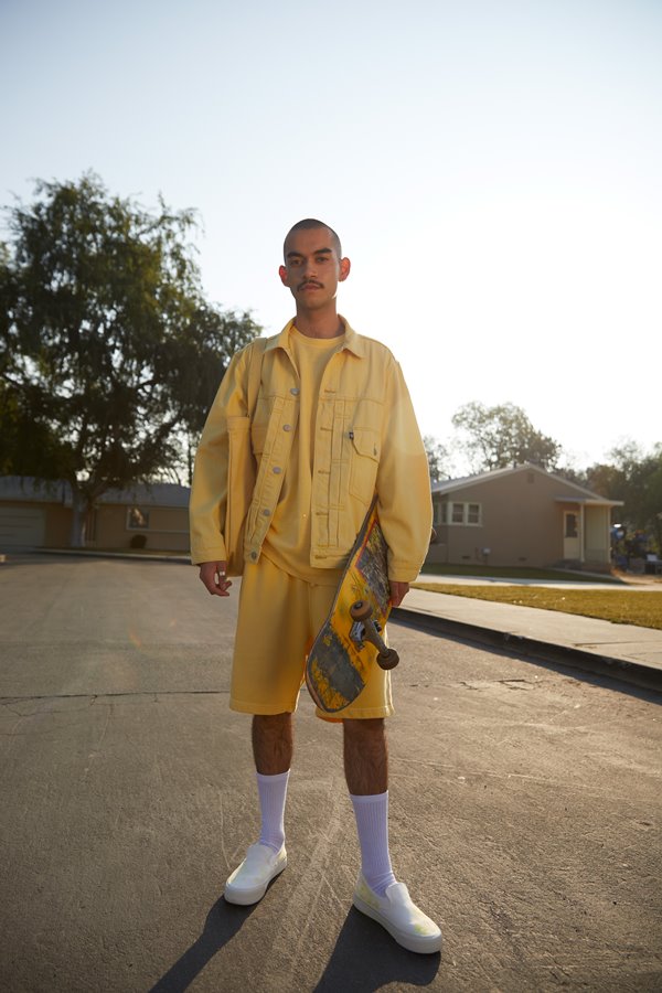 Homem branco e com o cabelo raspado e de bigode segurando um skate amarelo. Ele usa uma camiseta, jaqueta e shorts, também amarelos, meias brancas e tênis preto. As peças são da Levi's.