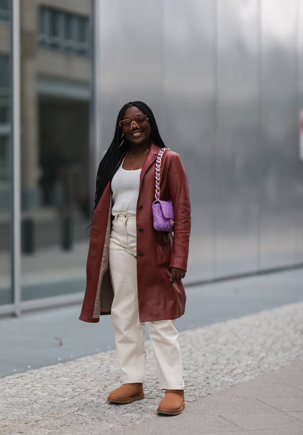 Mulher negra no street style usando look com calça bege de cintura alta, camiseta branca, sobretudo de couro vinho e bolsa baguete roxa