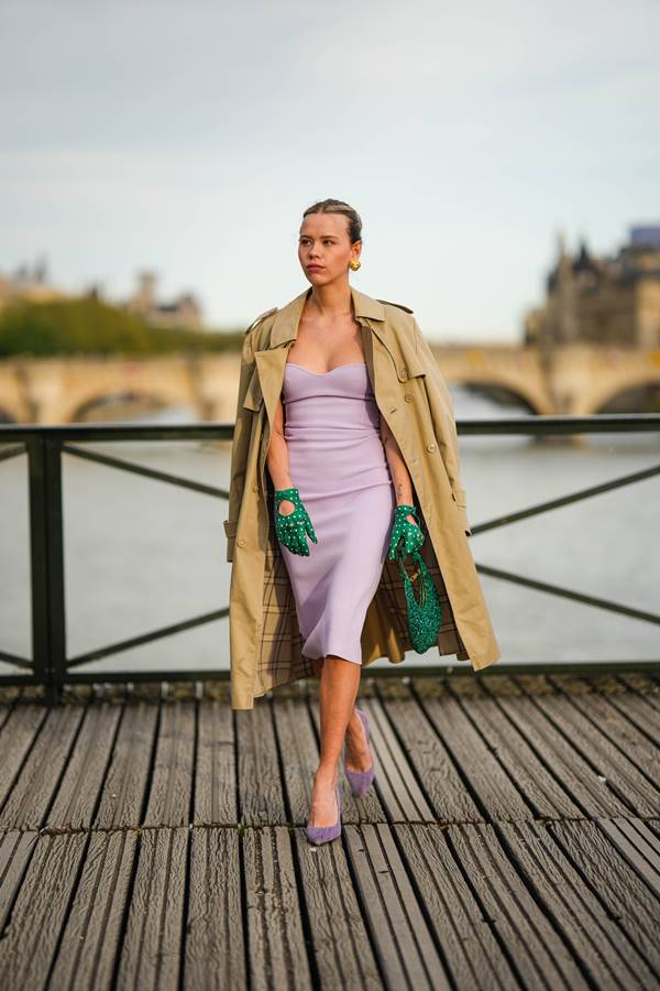 Mulher branca no street style usando vestido e sandália de salto lilases, com trench coat bege, além de luvas e bolsa verdes