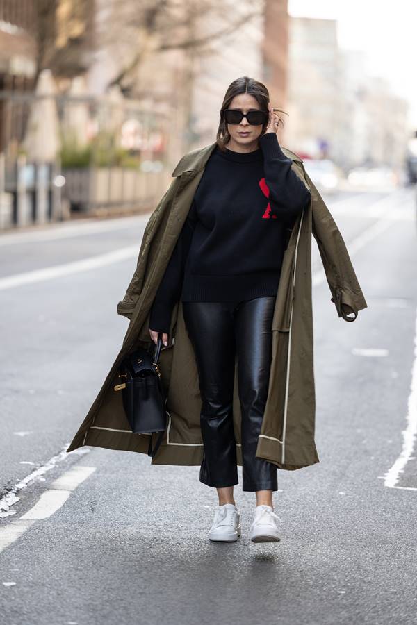 Mulher branca no street style usando calça preta de couro, moletom azul escuro, tênis branco, bolsa preta e por cima dos ombros trench coat verde musgo