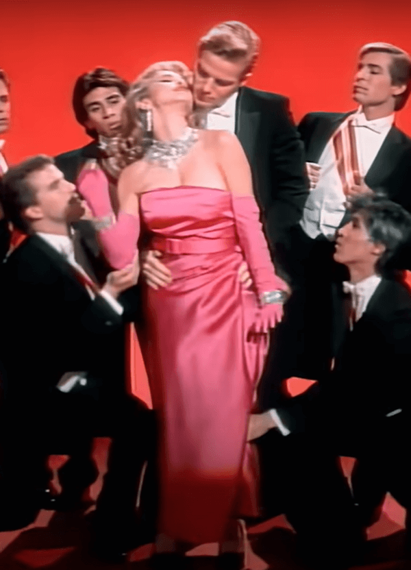 Na imagem com cor, Madonna está ao centro, usando um longo vestido rosa, enquanto homens usando ternos pretos a cercam 
