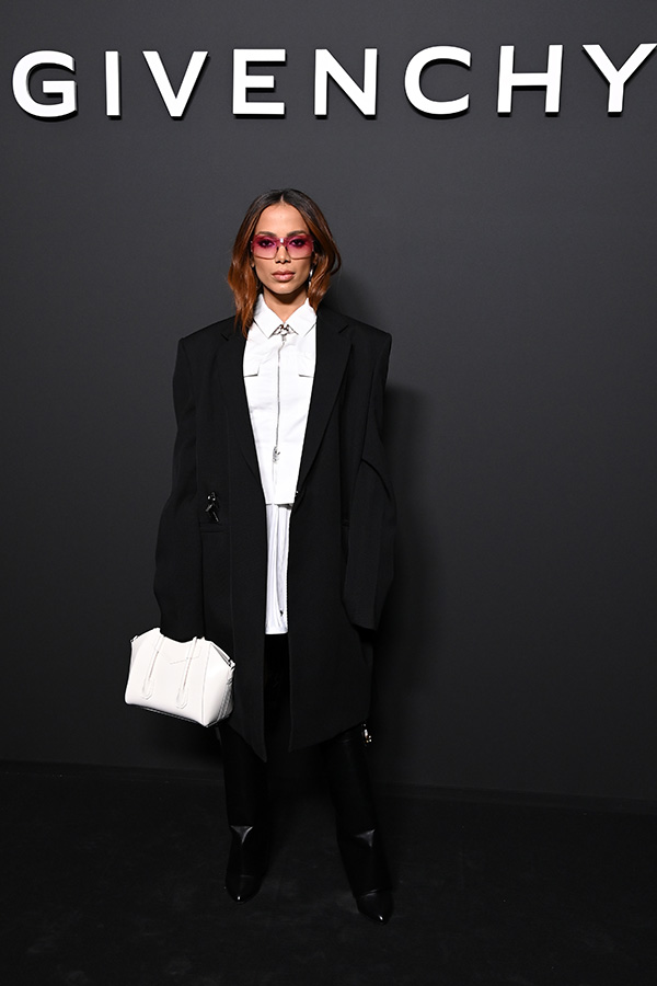 Cantora Anitta na porta do desfile da marca Givenchy. Ela usa uma camisa branca, calça e blazer preto, uma bolsa branca e óculos escuros cor de rosa.