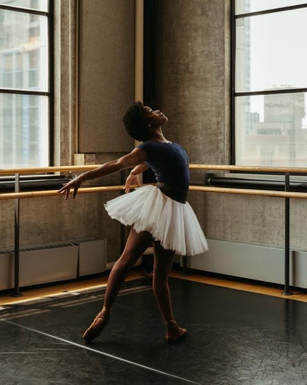 Mulher negra com maiô, saia de tule e sapatilhas de balé. Na sala, é possível ver barras para alongamento. O nome da bailarina é Ingrid Silva.