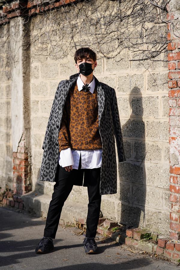 Homem branco com cabelo preto, curto e liso. Ele usa uma máscara preta, um casaco cinza com estampa de leopardo, um pulôver marrom de lã com estampa de leopardo, uma camisa branca, calça preta, tênis.