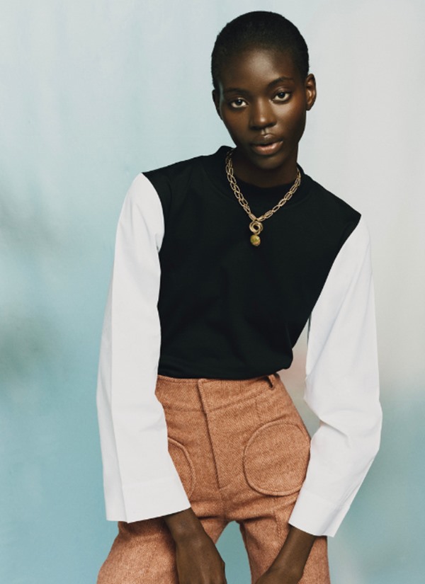 Mulher negra com cabelo amarrados posando para foto. Ela usa uma camisa branca, um pullover preto e uma calça marrom. Por cima das peças é possível ver um colar dourado.