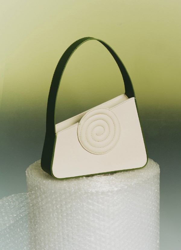 Bolsa branca com alça e extremidades em verde escuro. A peça possui ainda um detalhe de aspiral branco. Ela está repousada em um rolo de plástico bolha. O fundo da foto é um degradê de verde.