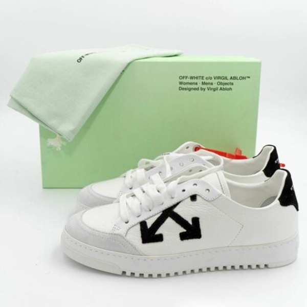 Tênis branco com detalhes pretos da marca Off-White. Atrás do tênis tem uma caixa verdinha