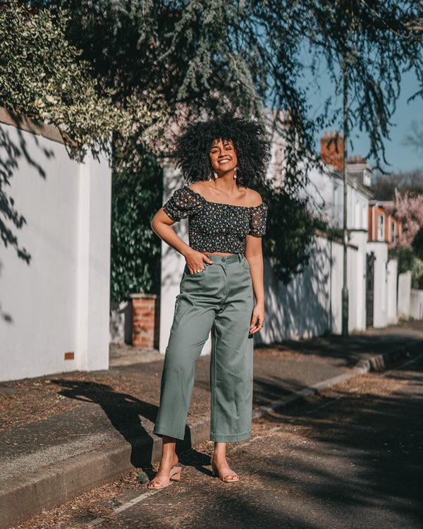 Mulher negra com cabelo cacheado posando para foto em uma rua arborizada. Ela usa uma blusa cropped e uma calça verde