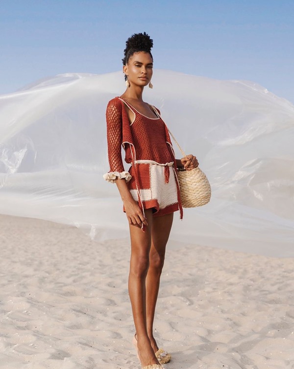 Mulher negra com cabelo cacheado amarrado em um coque posando para foto na praia. Ela usa saia, top e casaco, tudo de crochê marrom. As roupas são da marca Ateliê Mão de Mãe. - Metrópoles