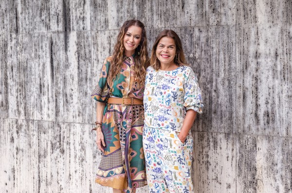 Duas mulheres brancas posando para foto. A primeira possui os cabelos longos e ondulados, em um tom de caramelo. A segunda tem o cabelo liso e curto. Ambas vestem roupas estampadas