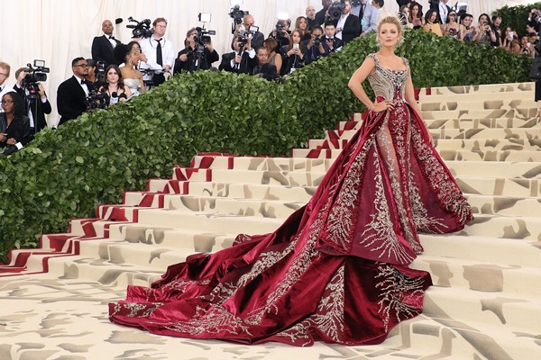 Mulher branca e loura em escadas da entrada de evento de moda. Ela usa um vestido vermelho e dourado com uma saia gigante. Seu nome é Blake Lively.