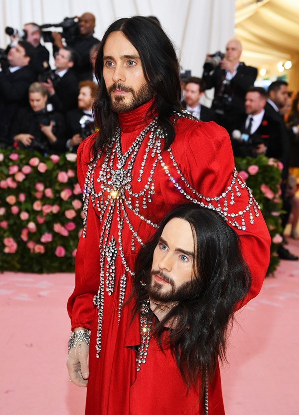 O ator e cantor Jared Leto com um vestido vermelho e correntes pratas por cima. Ele segura uma cabeça cenográfica que imita seu próprio rosto. Ele está no tapete vermelho do baile do Met Gala, evento da revista Vogue.