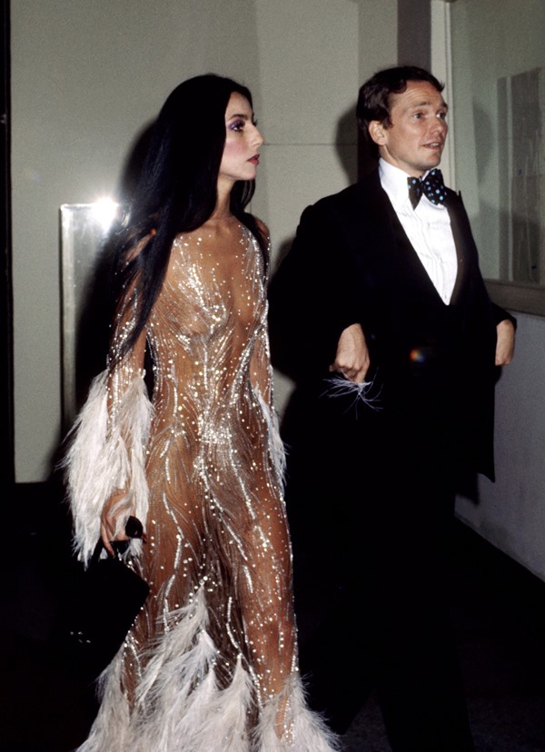 A cantora Cher, em 1974, chegando no Met Gala com o estilista Bob Mackie. Ela usa um vestido de tule transparente com faixas de glitter e mangas de pluma. Ele usa um smoking clássico, com blazer e calça pretos e camisa branca.