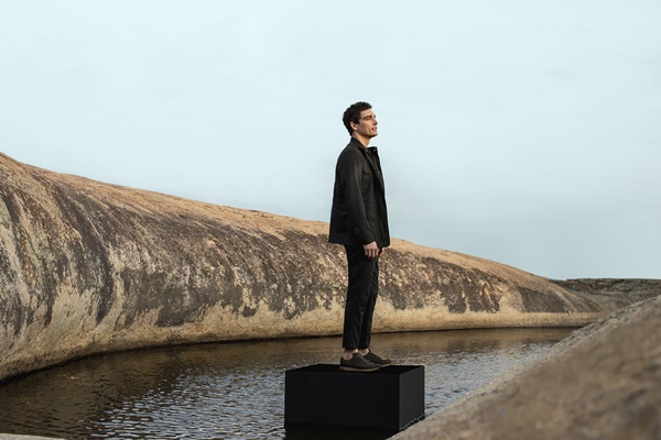 Homem branco com cabelo preto vestindo roupas pretas e posando em um lago, no cariri paraibano. 