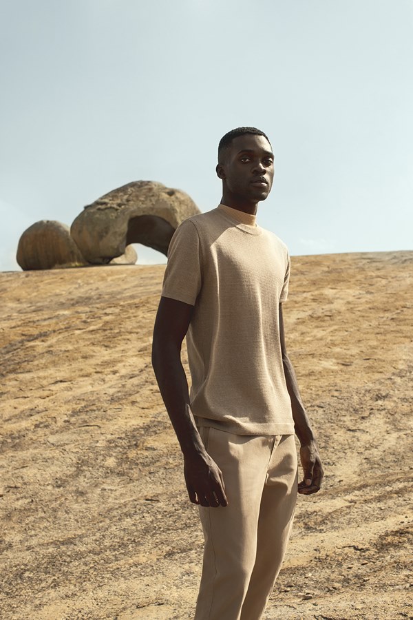 Homem negro, com cabeça raspada, vestindo camiseta e calça bege. Ele está na natureza, na região do cariri paraibano, com solo marrom e pedras no mesmo tom 